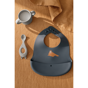 Stylish gray silicone bib with a playful penguin design, accompanied by a soft feeding spoon on a natural cloth backdrop.