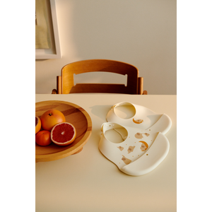 Two soft silicone bibs in playful designs beside a wooden bowl of oranges on a bright table.