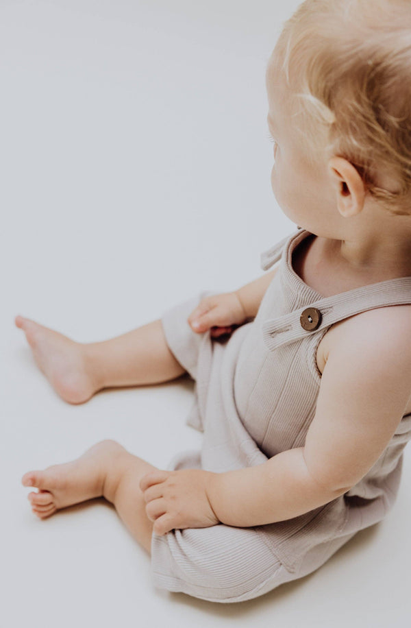 Kleines Kind sitzt in einer Latzhose aus weichem Rippstrick in Stone, ideal für den Sommer.