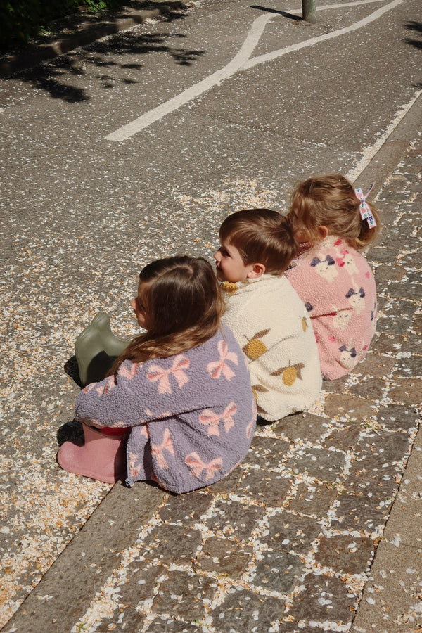 Three children sitting on a sunny pavement wearing cozy sweaters, surrounded by flower petals.