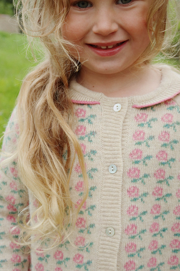 Sweet girl wearing a floral knit cardigan with pink roses, showcasing buttons and cozy design in a natural outdoor setting.