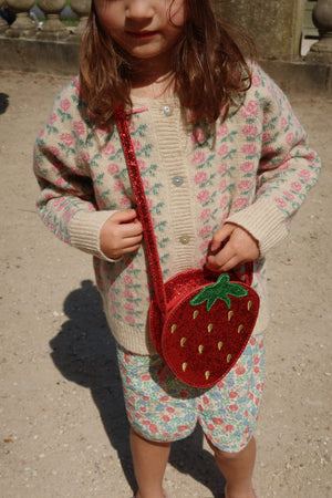 Child wearing a soft cardigan and a strawberry-shaped purse, enjoying a sunny day outdoors.