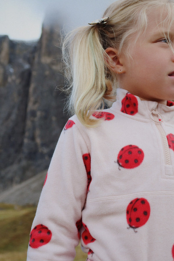 A child wearing a cozy Tavi Fleece-Sweatshirt with ladybug prints, enjoying the outdoors in a scenic landscape.