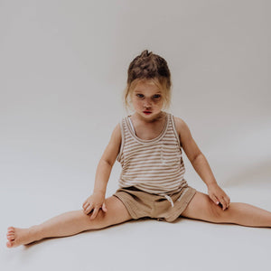 Ein kleines Mädchen in gestreiftem Oberteil und bronzefarbenen Musselin-Babyshorts, sitzt spielerisch auf dem Boden.